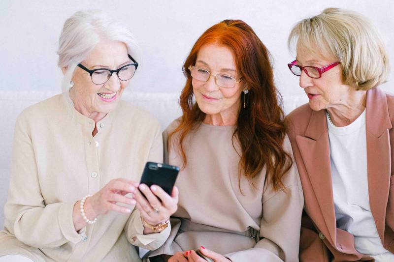 Trois femmes attentives consultent les données des montants moyens de retraite par département en France sur un téléphone. Cette photo illustre l'importance de comprendre les réalités financières des retraités français à travers les disparités régionales.